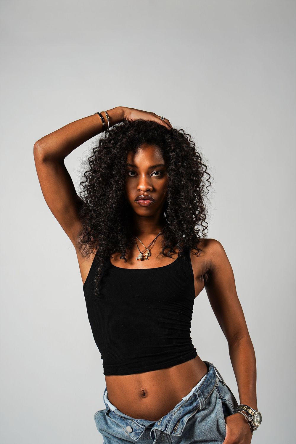 A person in a black crop top and jeans poses confidently in a studio against a gray background.