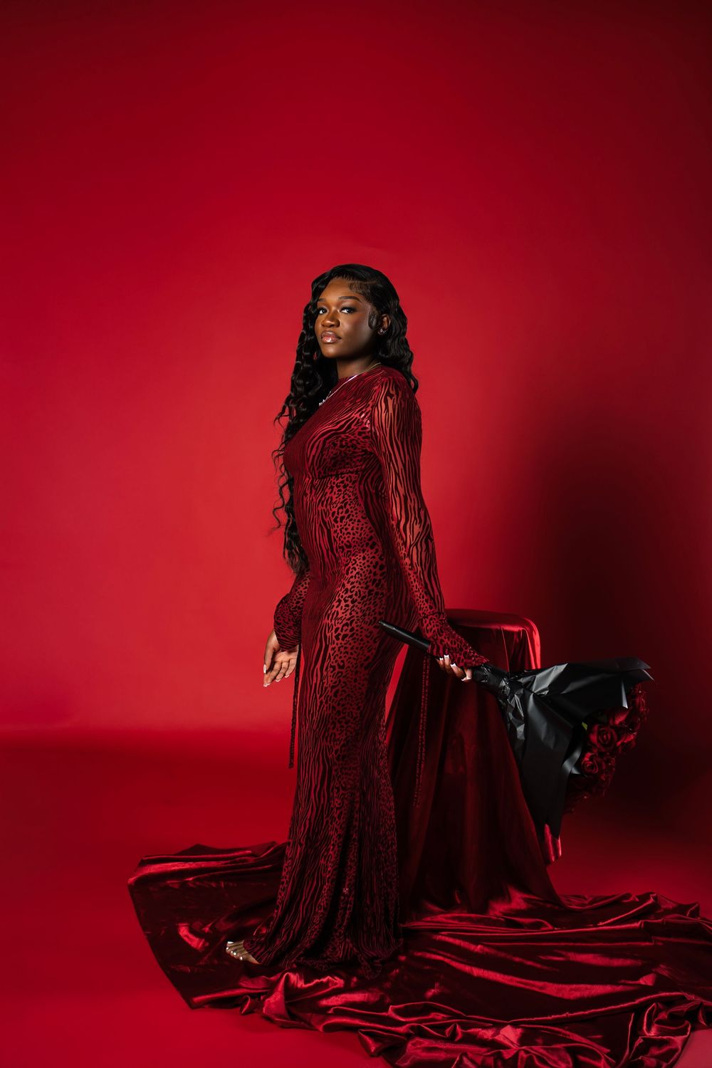 A figure in a flowing red velvet gown poses dramatically against a deep crimson backdrop in a moody photoshoot.