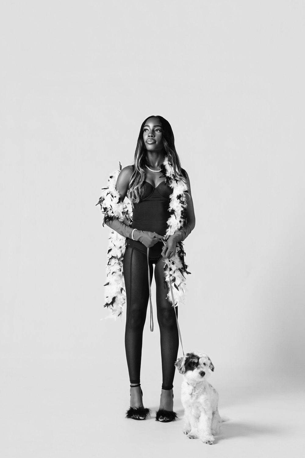 A figure in a black outfit poses with a fluffy white dog against a white background in a studio photoshoot series.