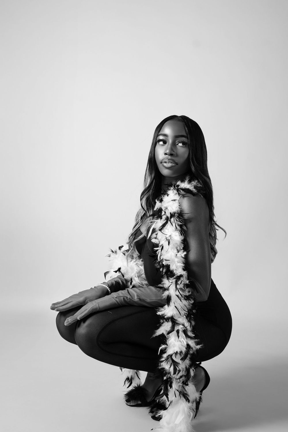 Black and white studio portrait of a model in a crouching pose wearing a feather boa against a plain background.