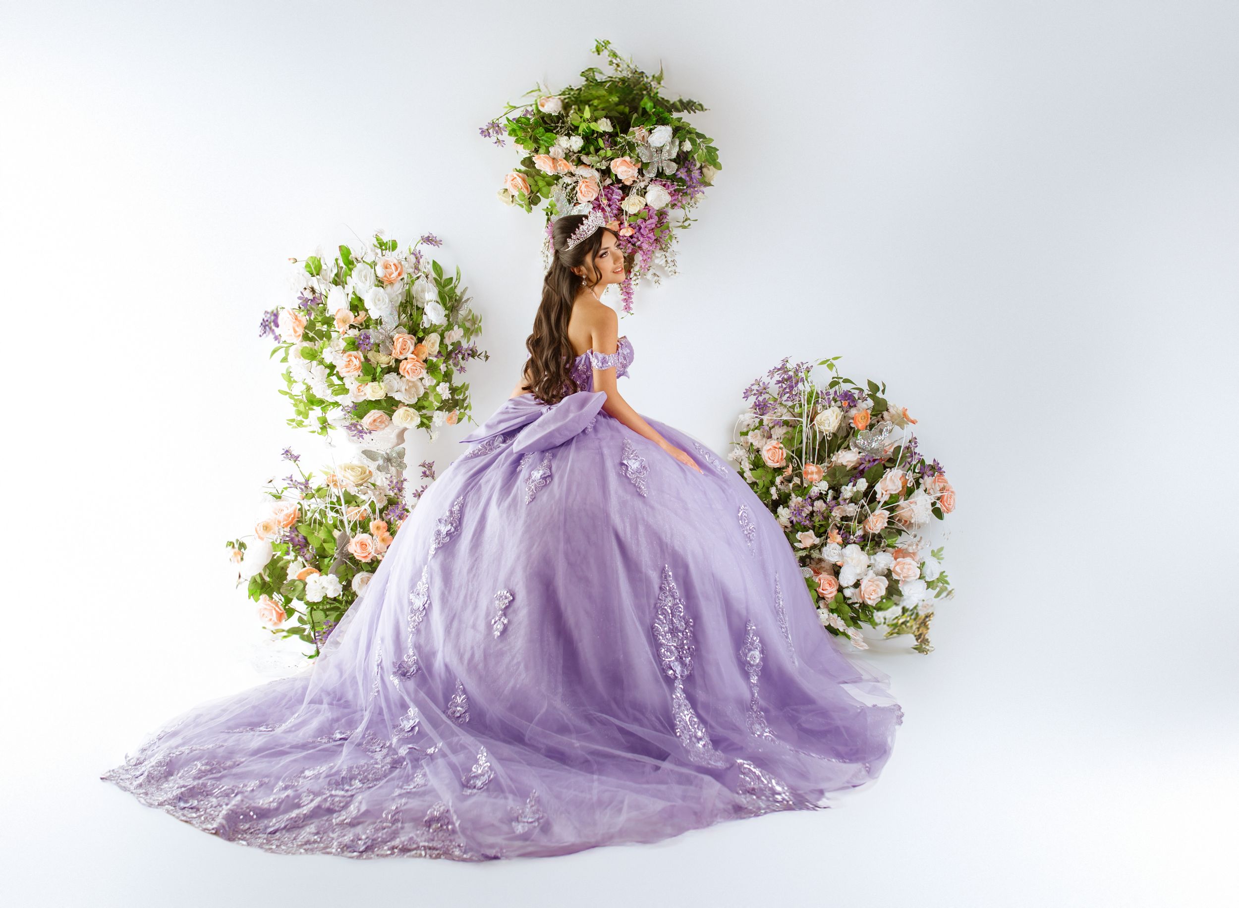 A lavender tulle ballgown with floral embellishments photographed against white backdrop with floral arrangements.