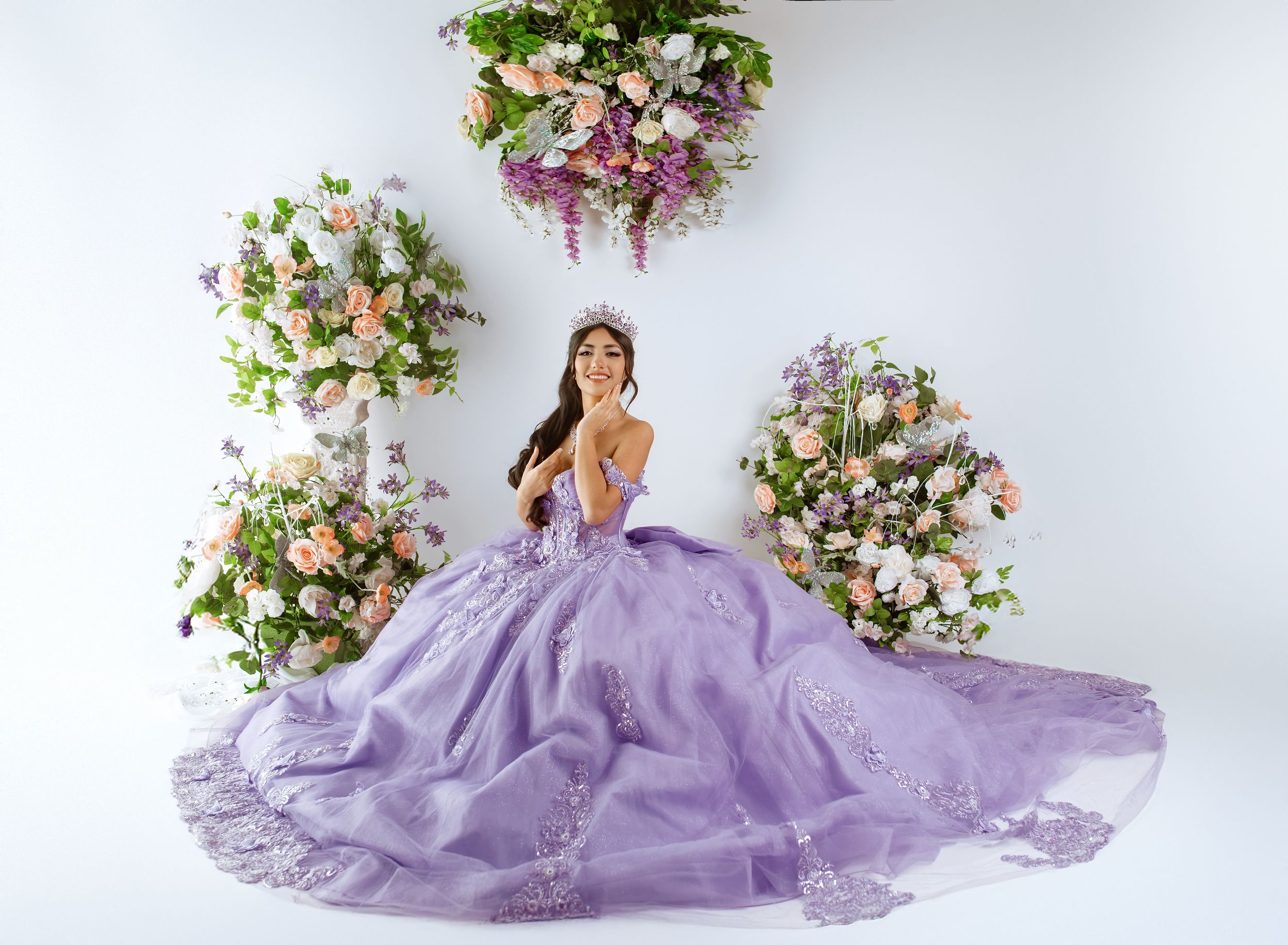 A model in a flowing purple tulle ball gown poses elegantly against a white backdrop with floral arrangements.