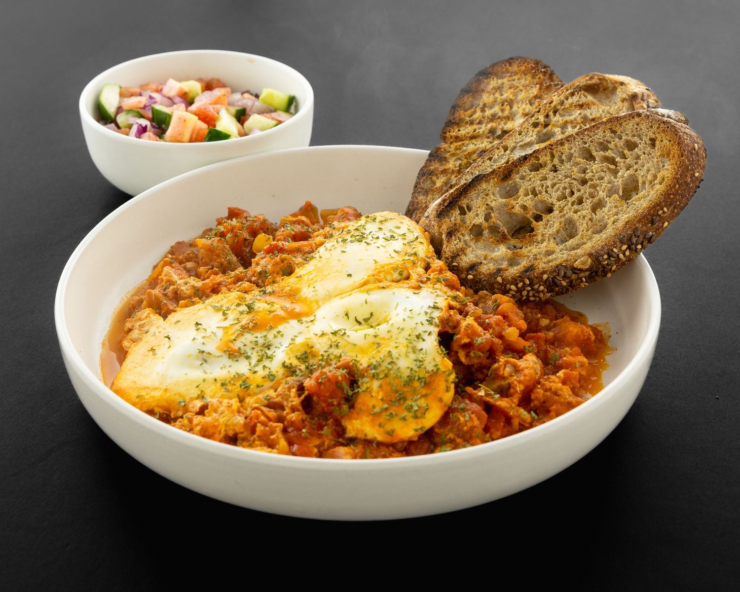 A steaming bowl of shakshuka with eggs, tomatoes and bread served on a dark background.