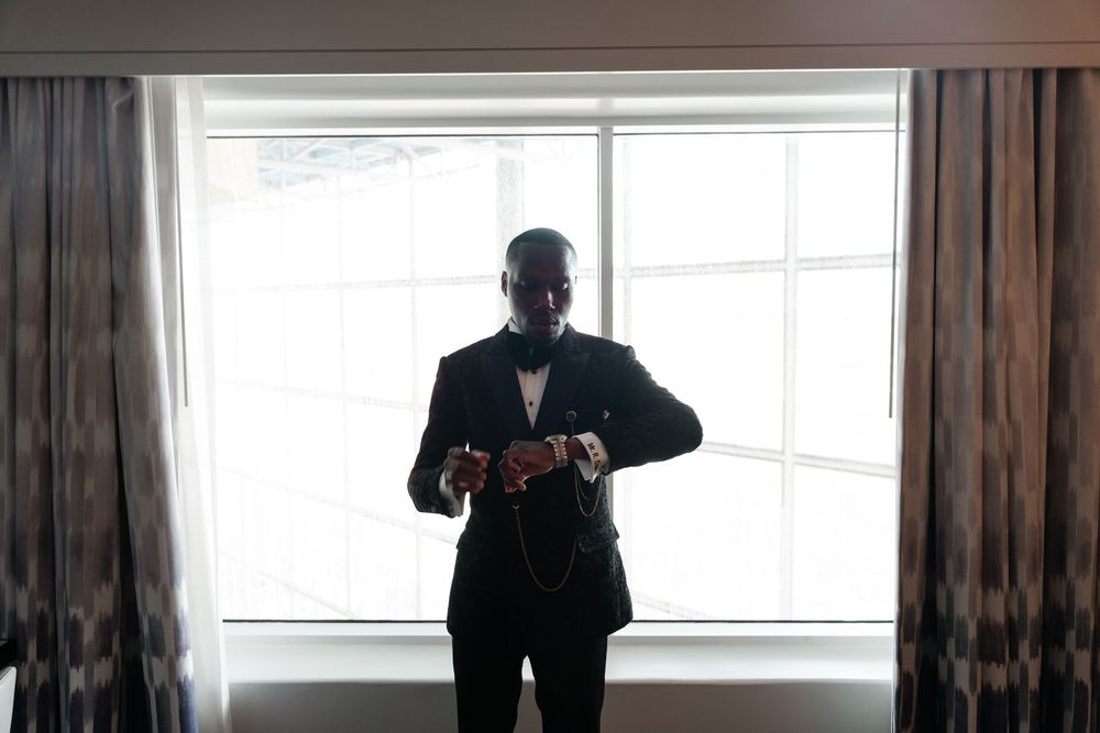A person in a dark suit stands by a window in a dimly lit room with curtains.