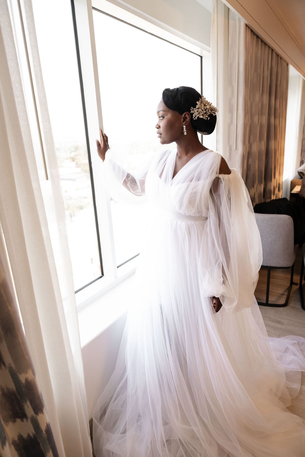 A bride in a flowing white wedding gown and veil stands elegantly by sunlit windows in a romantic bridal portrait series.