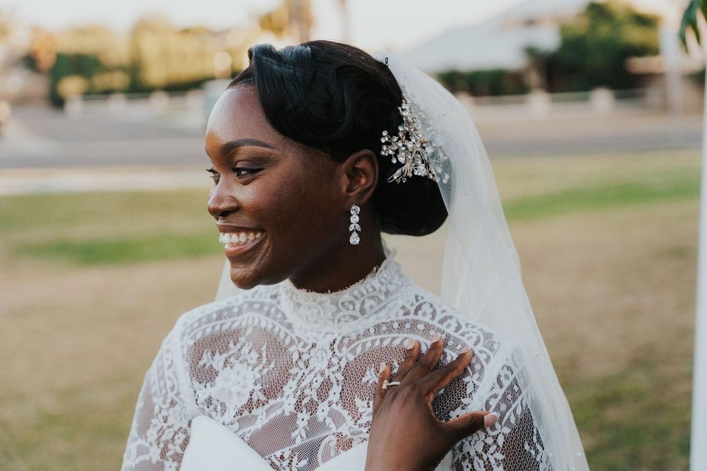 A bride wearing an elegant lace wedding dress with a veil and delicate floral hair accessories smiles outdoors.