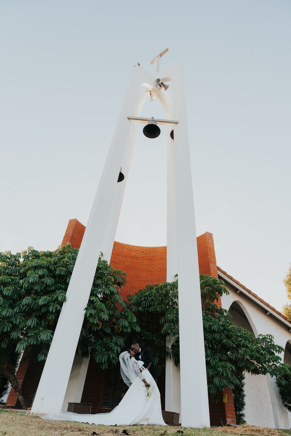 A couple stands beneath a tall white and orange architectural tower structure with trees in the background at sunset.