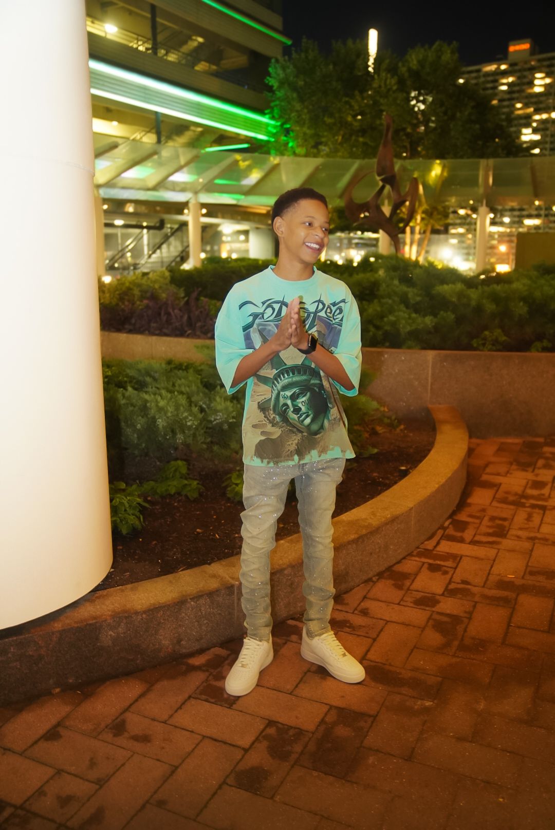A figure stands on brick pavers under green lighting near illuminated columns at night.