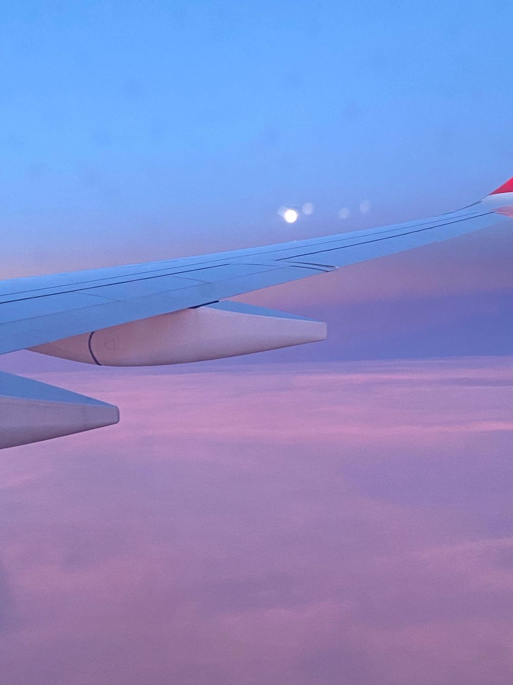 View of airplane wing against dreamy pink and purple sunset sky with full moon visible in the distance.