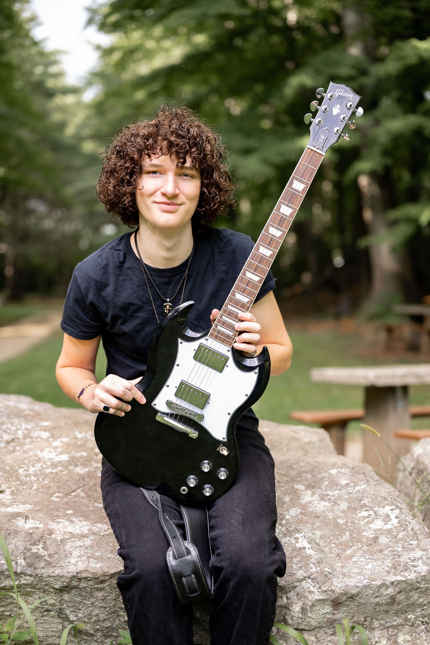 A musician in black clothing sits on a stone wall outdoors while posing with an electric guitar.