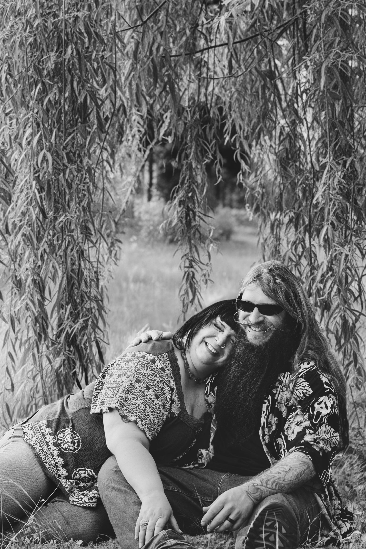 A couple relaxes together under the drooping branches of a willow tree in a black and white photograph.