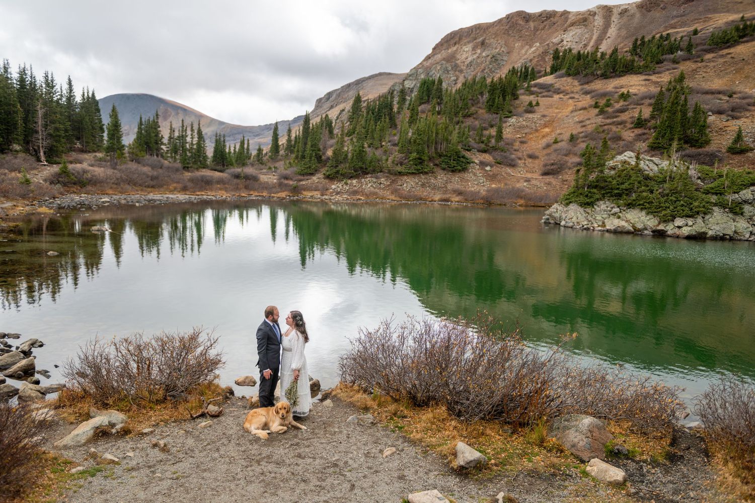 An eloping couple stands by a stunning alpine lake with emerald waters, surrounded by mountains and pine trees during an outdoor elopement.