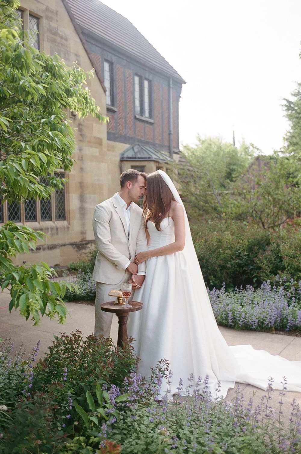 A couple shares an intimate moment in a garden with blooming lavender flowers and stone architecture in the background.