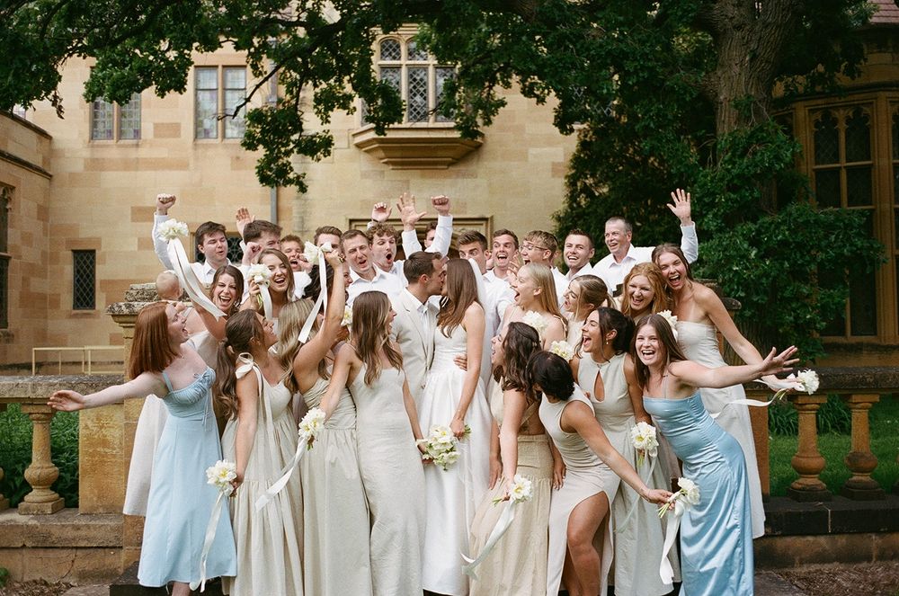 Wedding party in light blue and cream dresses celebrates joyfully on stone steps of historic building.