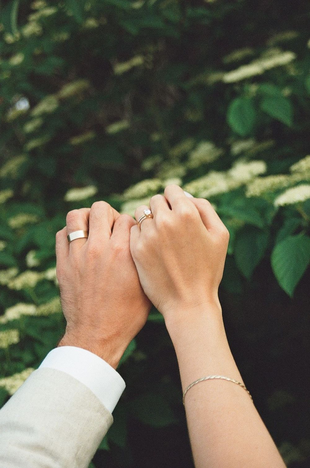 Close-up of two hands wearing wedding rings and jewelry against a green leafy background.