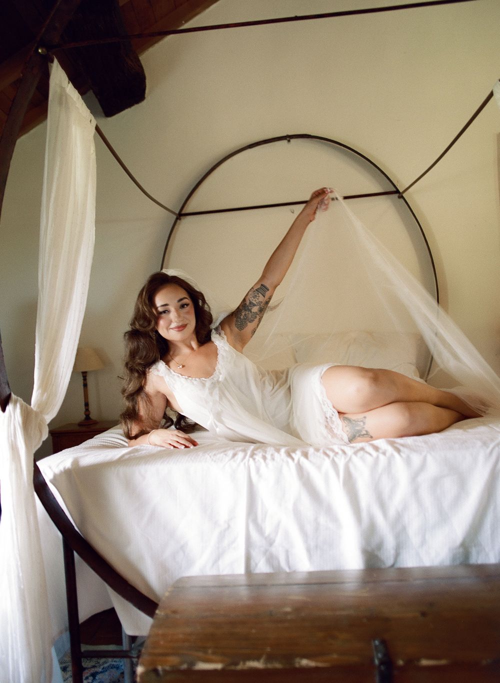 A tattooed brunette woman in white lingerie poses gracefully on a canopy bed with sheer draping.