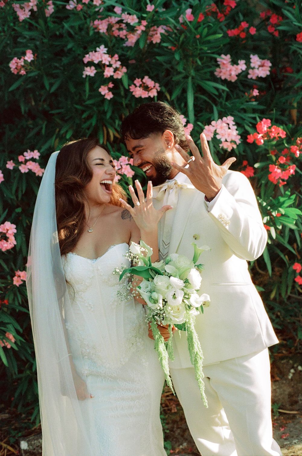Happy newlyweds showing wedding rings, bride in white gown with bouquet, groom in cream suit, pink flowers background.