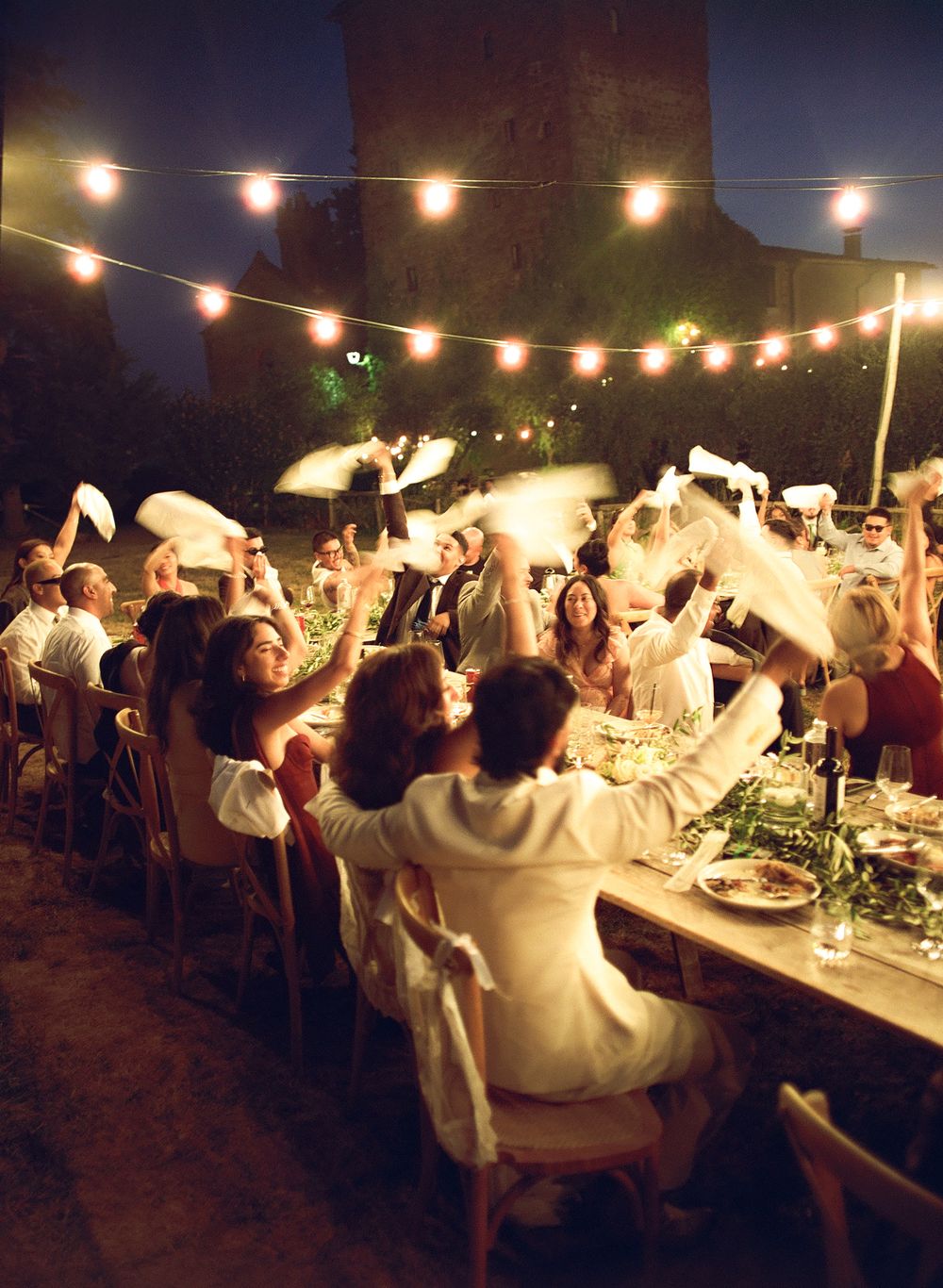 Guests wave napkins at an outdoor evening wedding reception with string lights and long dining tables.
