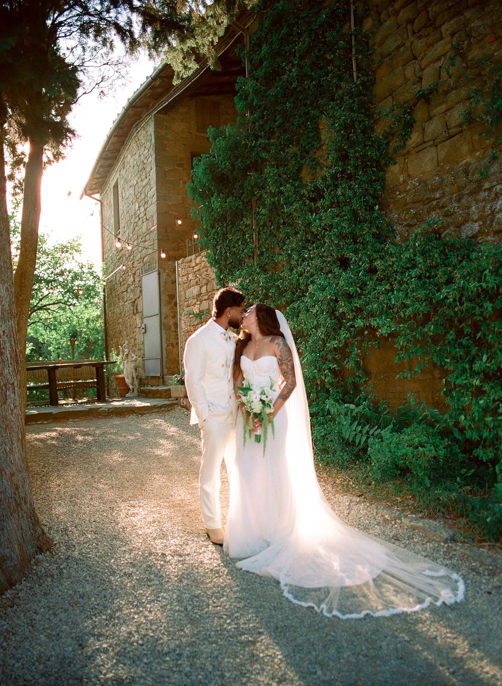 Bride and groom share a romantic kiss outside a stone villa surrounded by lush ivy in a sunlit Tuscan garden.