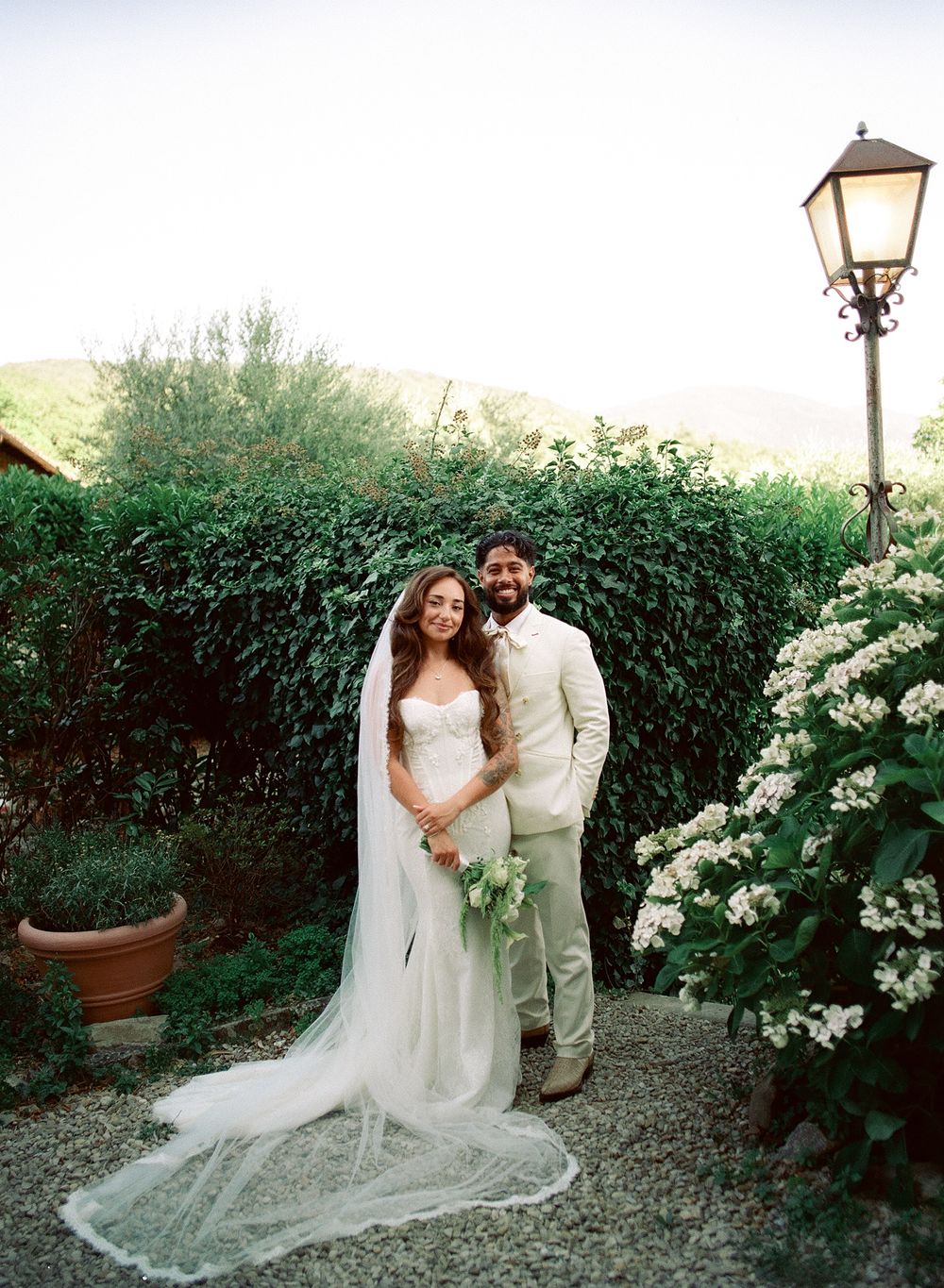 Bride in white gown with veil and groom in tan suit pose together in lush garden with flowering shrubs at outdoor wedding.