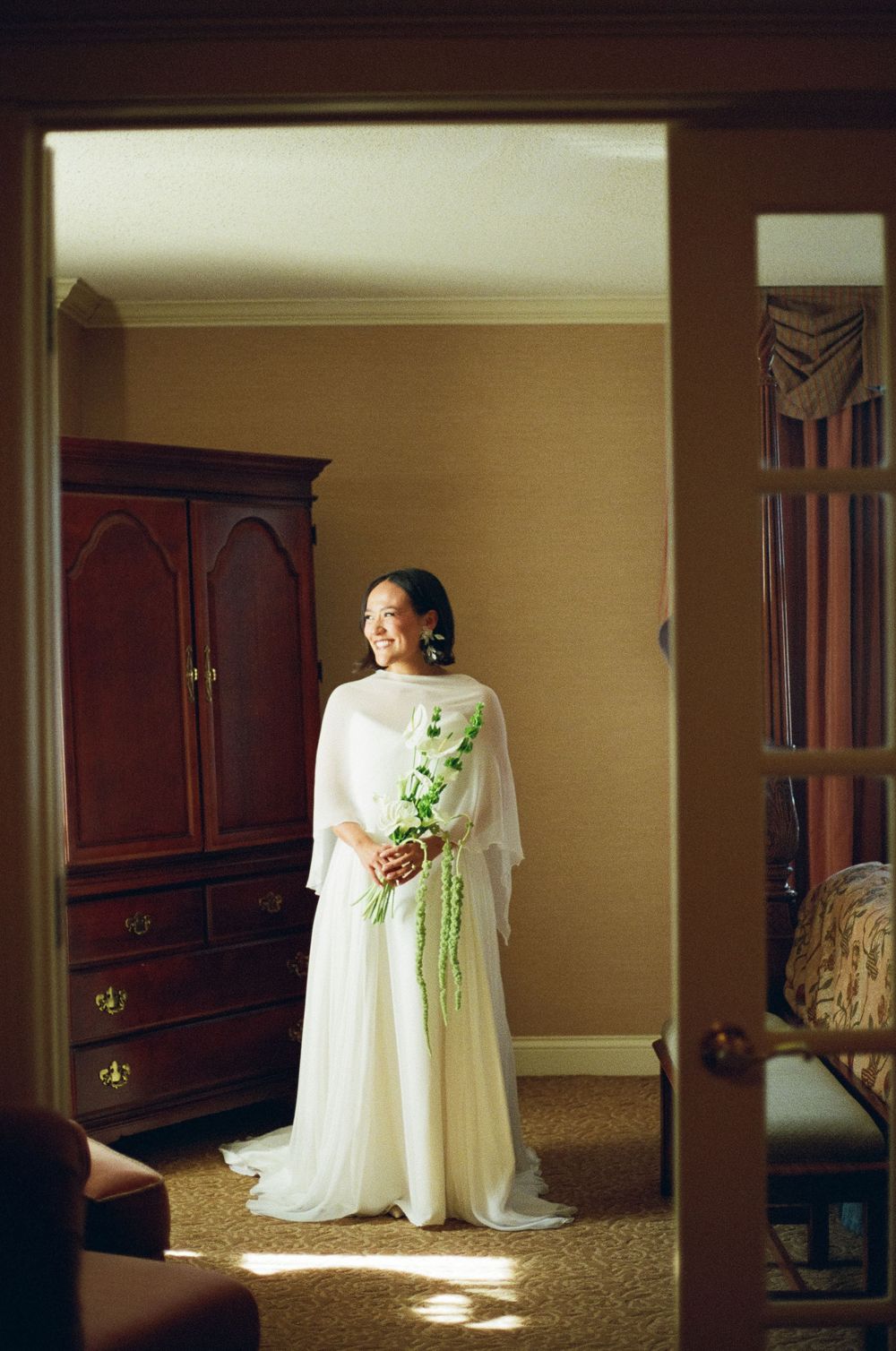Bride in white flowing gown with cape holds green bouquet, standing in sunlit room by wooden armoire.