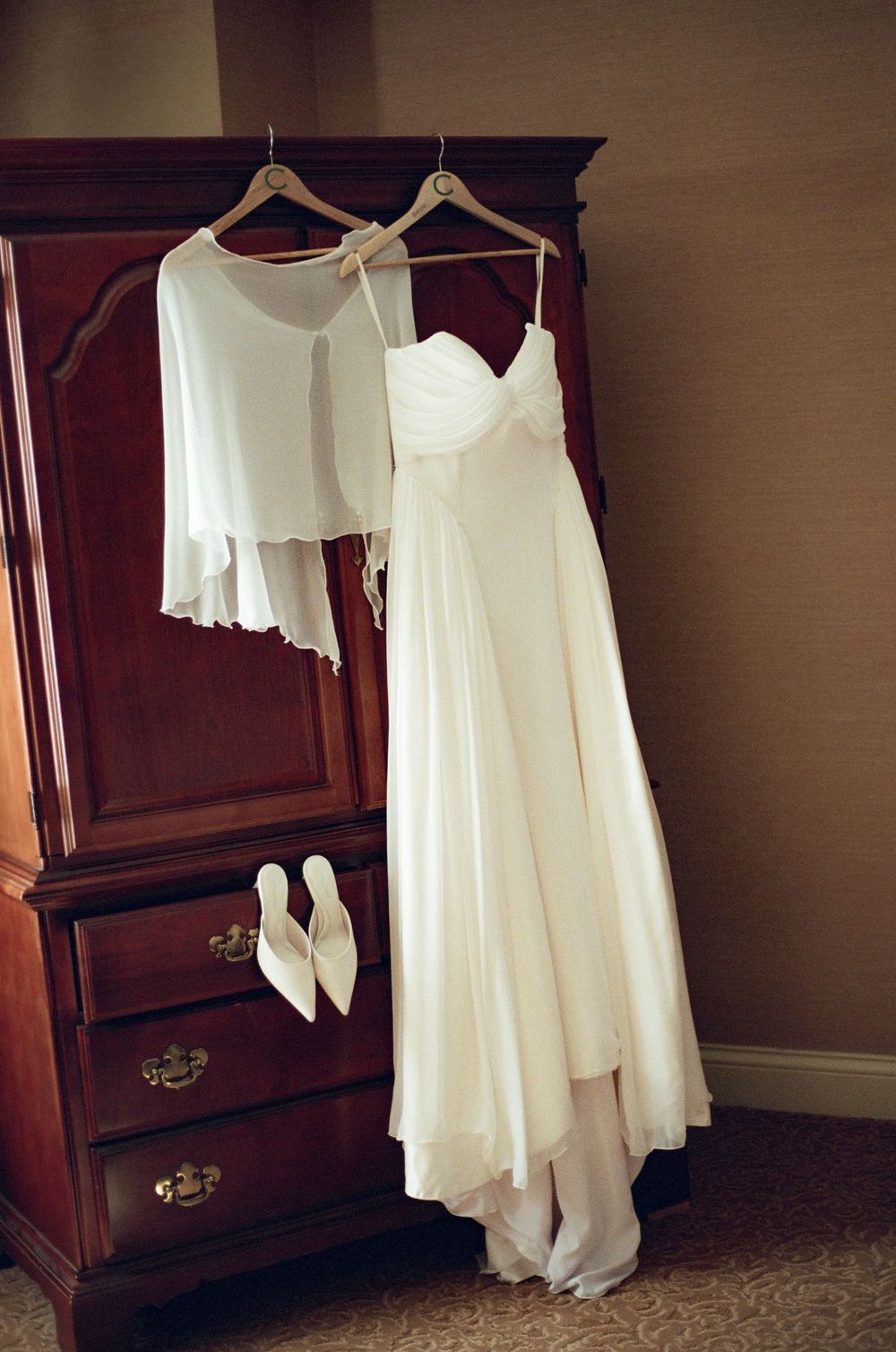 Elegant white wedding dress and shawl hanging on wooden armoire with matching bridal heels below.