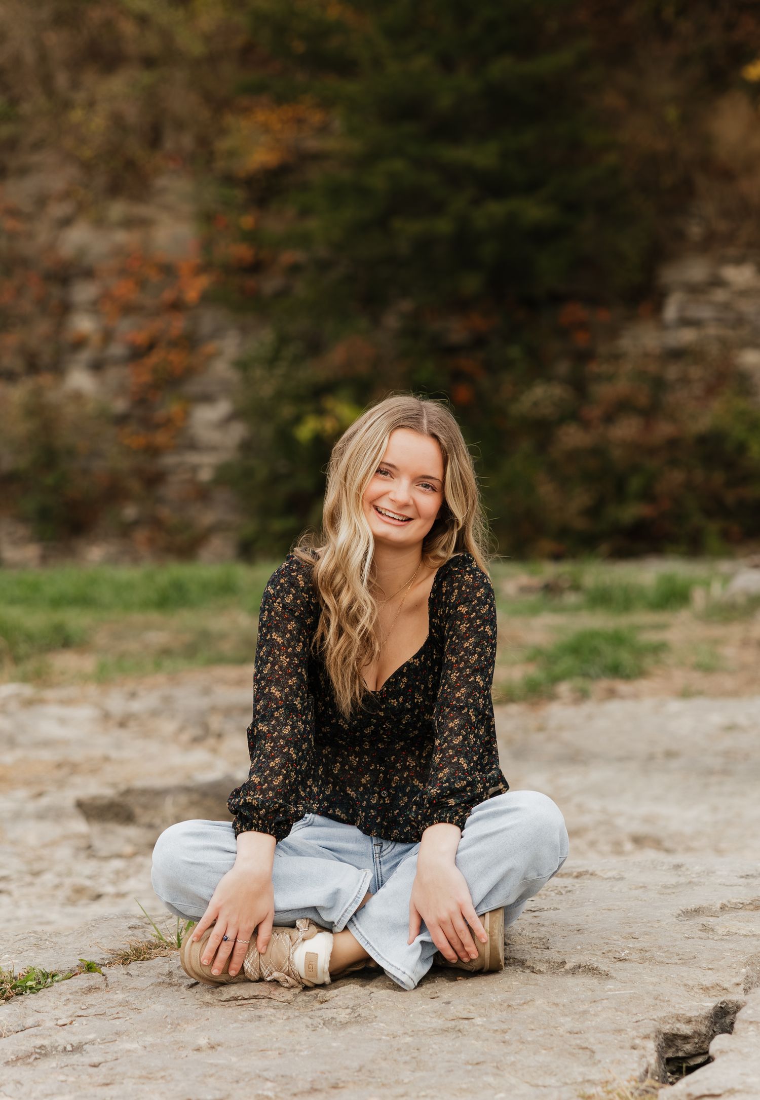 A person in a black floral top and light jeans sits cross-legged on the ground, smiling warmly on an autumn day.