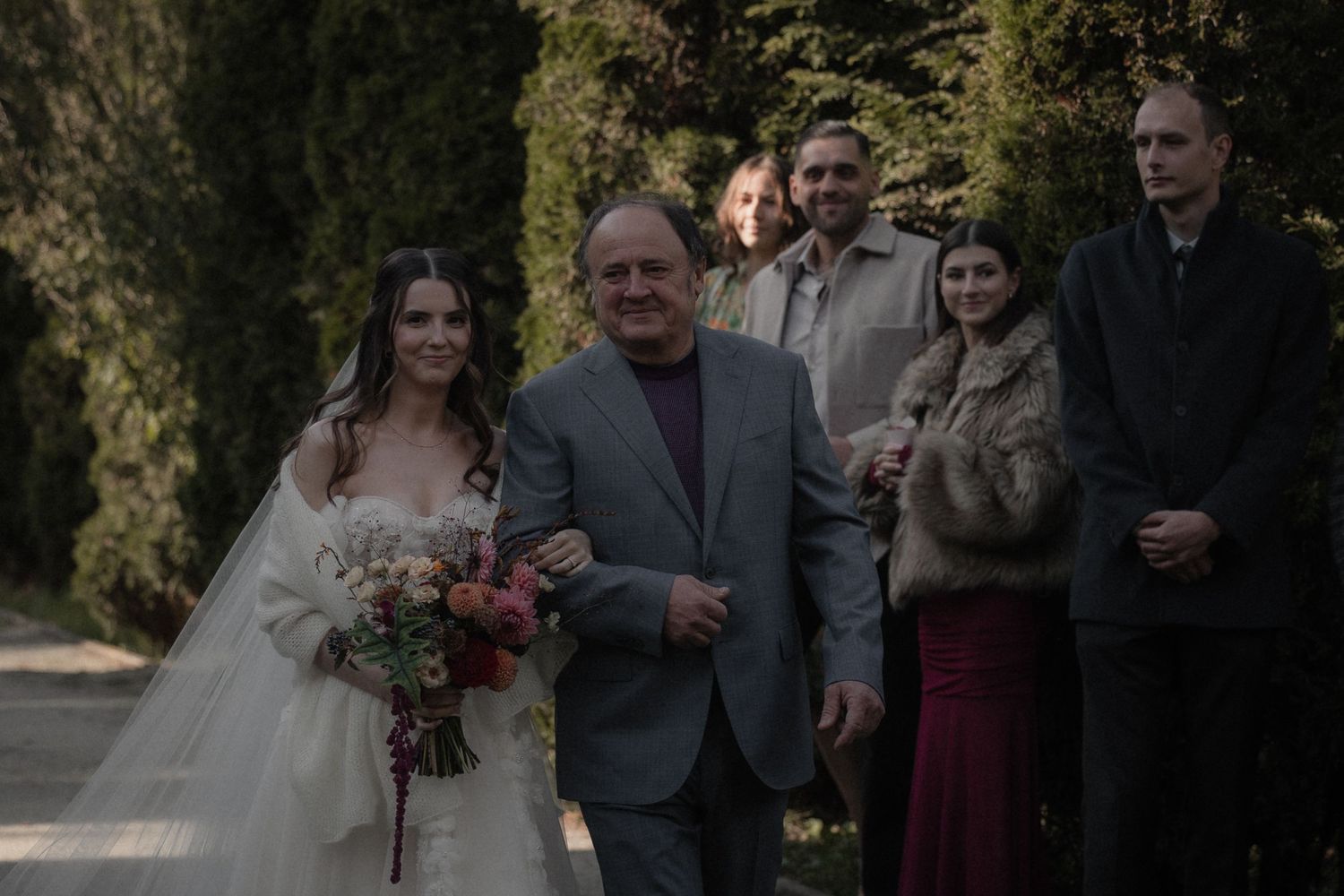 Wedding party walks together outdoors on a path surrounded by autumn trees and natural scenery.