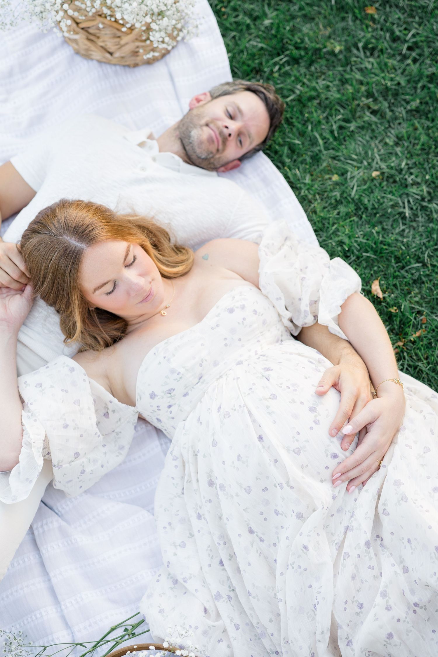 Couple in white wedding attire lying peacefully on green grass during outdoor photo session.