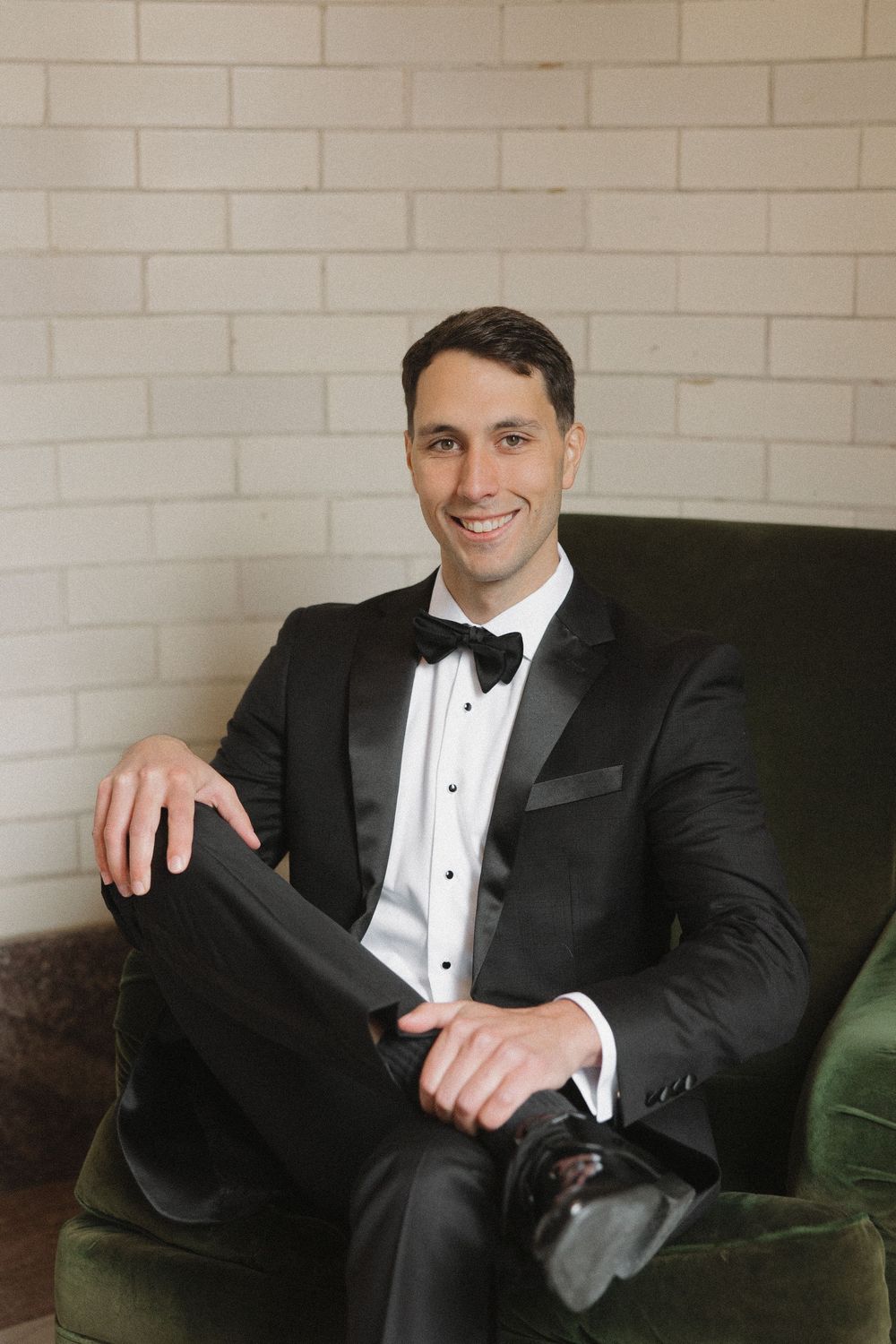 A person in a black tuxedo and bow tie sits relaxed on a green velvet couch against a white brick wall.