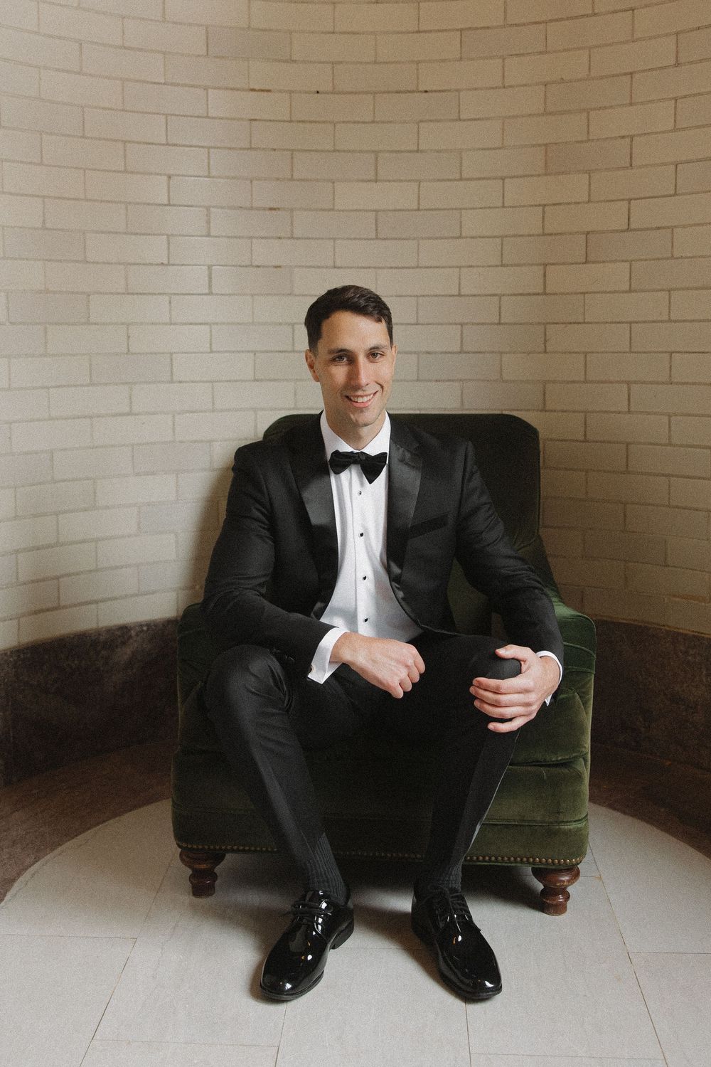 A person in a formal tuxedo and bow tie sits on a green velvet couch in a minimalist room with white tile and brick walls.