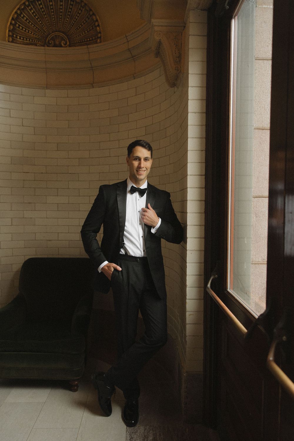 Groom in classic black tuxedo poses against architectural columns in elegant venue.