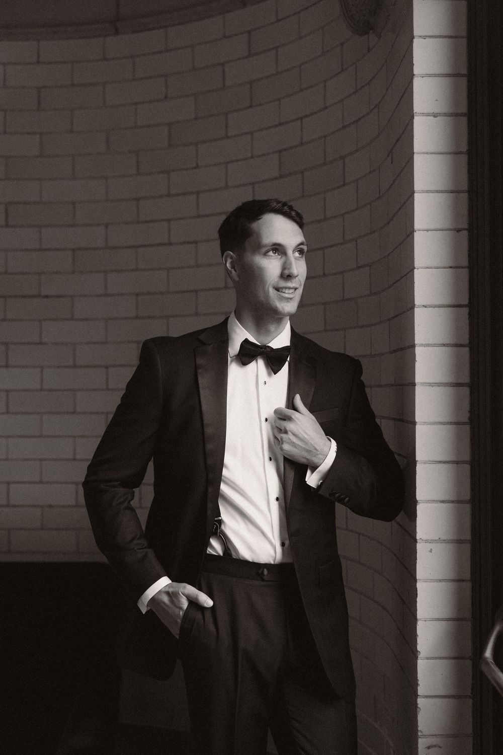 Classic black and white portrait of a person in a tuxedo standing against a brick wall with elegant lighting.