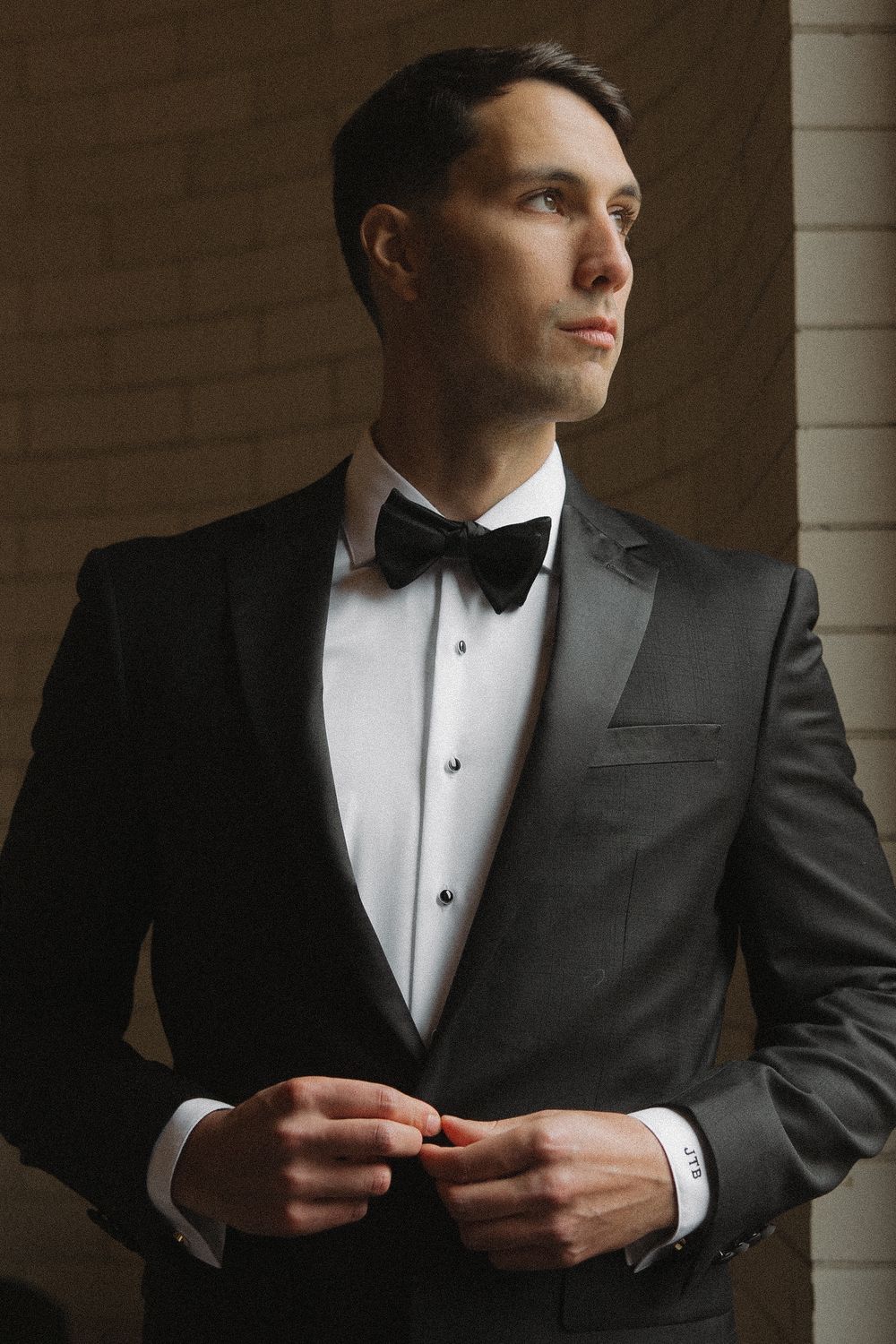 A groom in a classic black tuxedo and bow tie gazes thoughtfully to the side in an elegant portrait series.