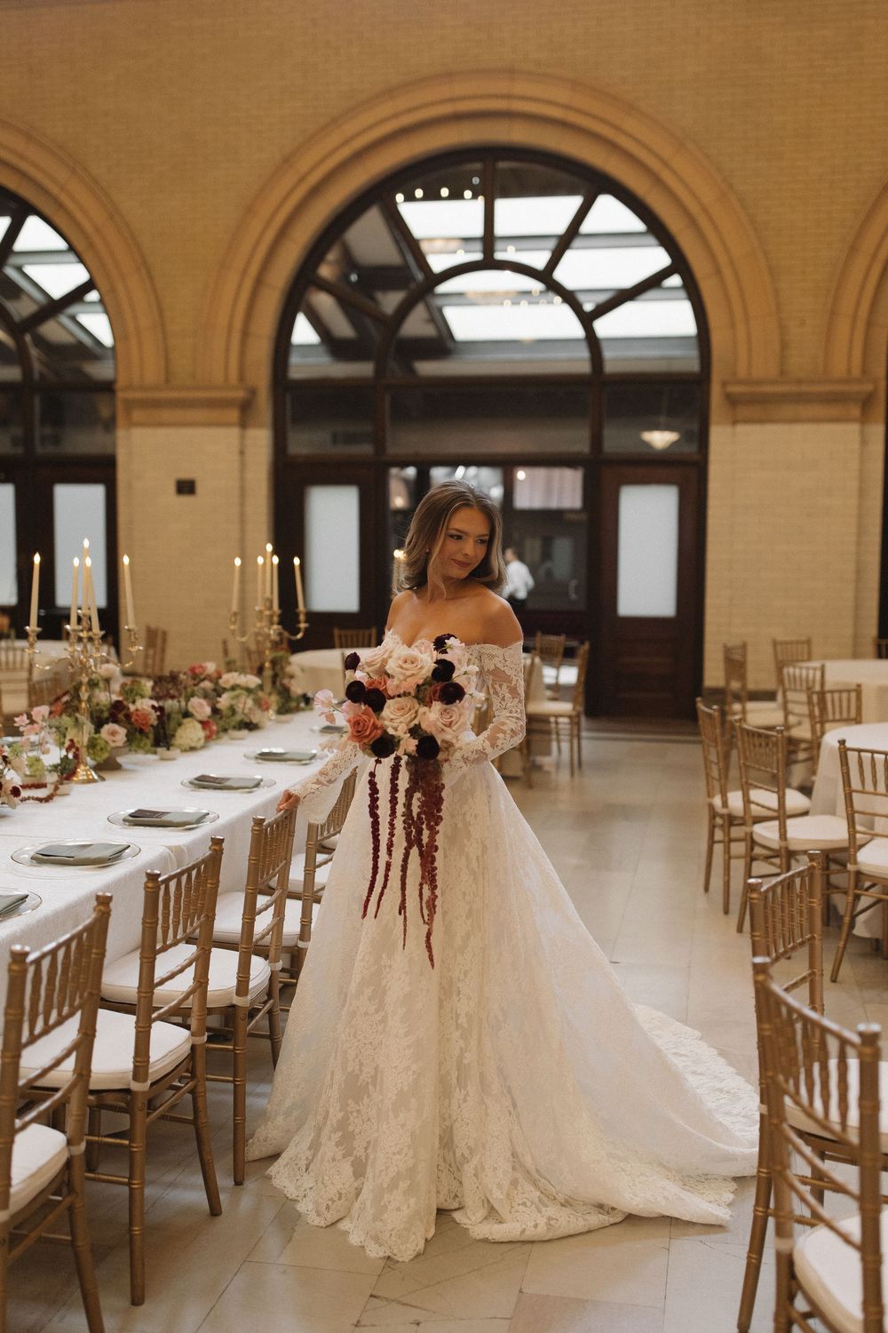 A bride in a lace off-shoulder wedding gown stands in an elegant venue with arched windows and long banquet tables.