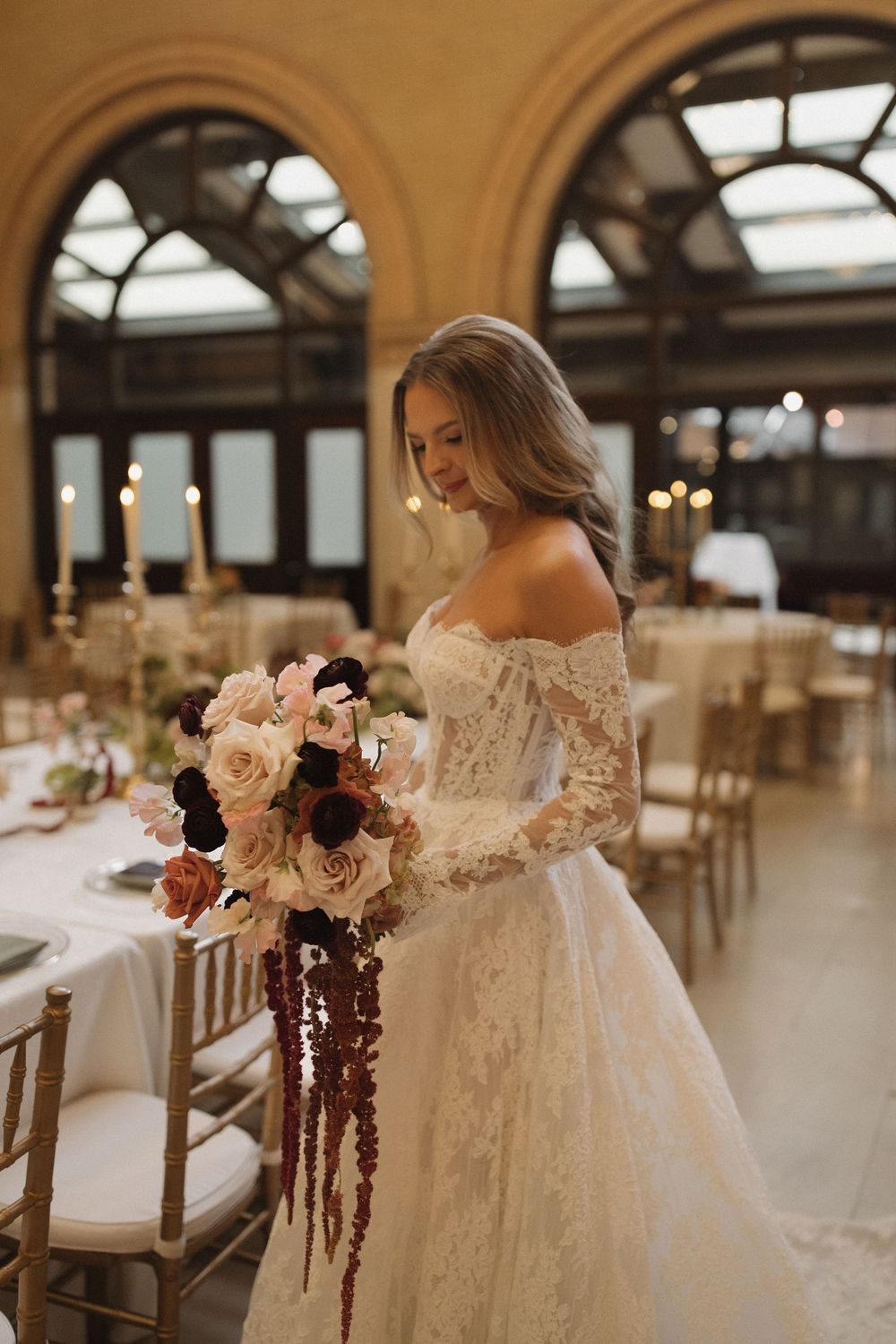 Bride in lace wedding gown stands beside floral arrangements in rustic reception venue.