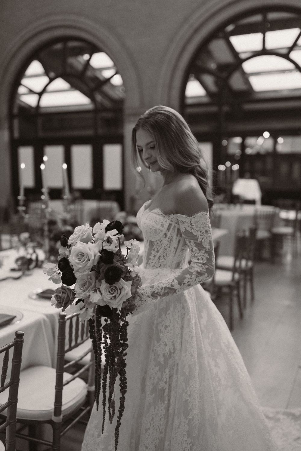 Elegant bride in a flowing white lace gown poses beside vintage wooden chairs in a sunlit venue.