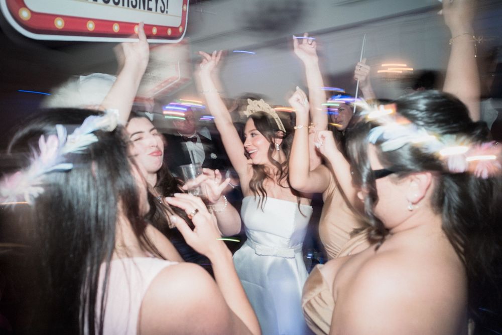 Bride and bridesmaids dancing joyfully at a wedding reception party with lights and raised hands.