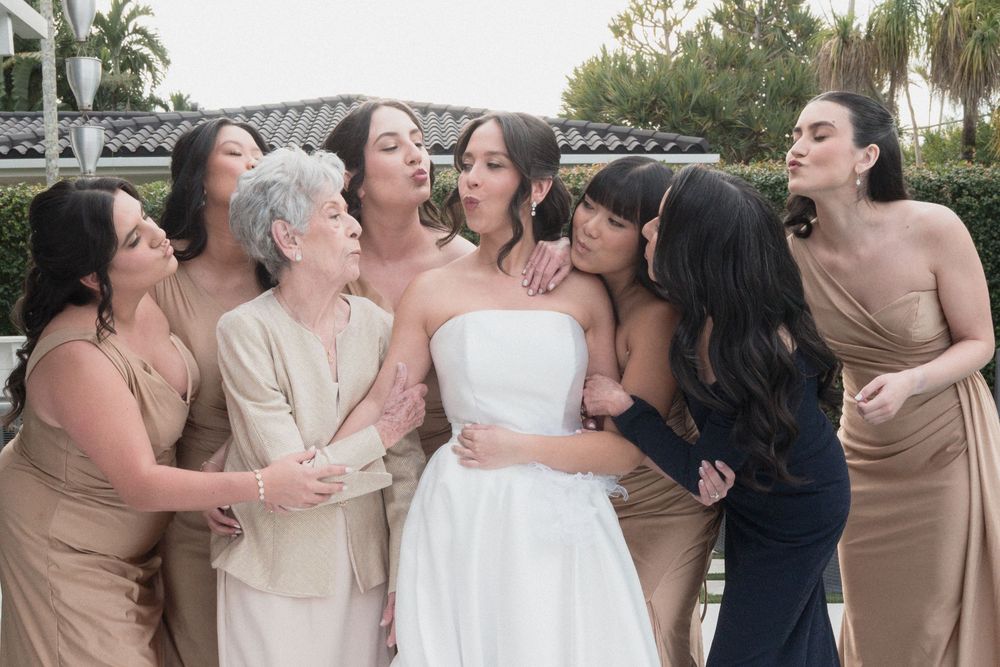 Bride in white strapless gown surrounded by bridesmaids in champagne and navy dresses sharing a joyful moment outdoors.