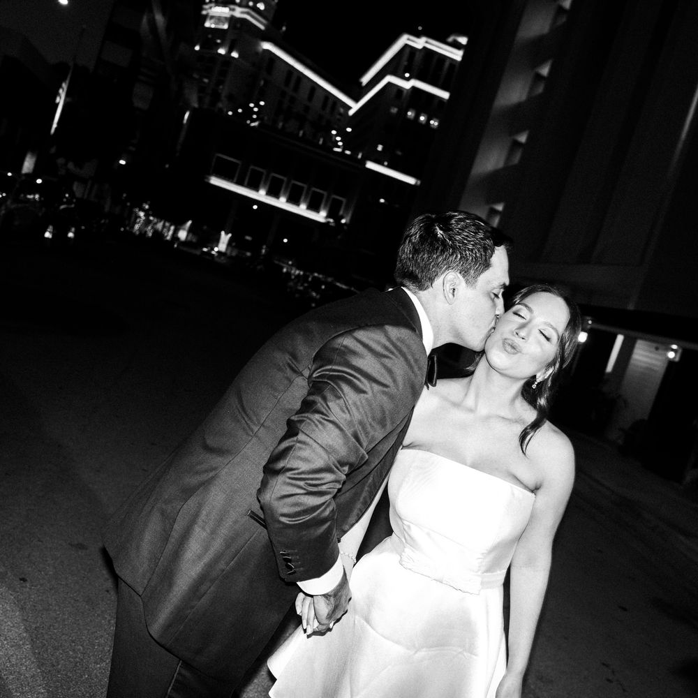 A romantic black and white nighttime photo of a couple in wedding attire sharing a kiss against a city skyline.