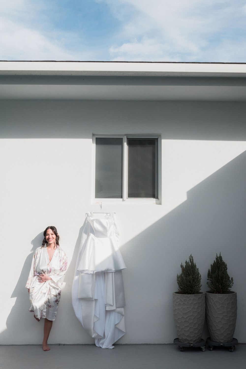 Bride in white robe stands beside hanging wedding dress against white wall with potted plants outdoors.