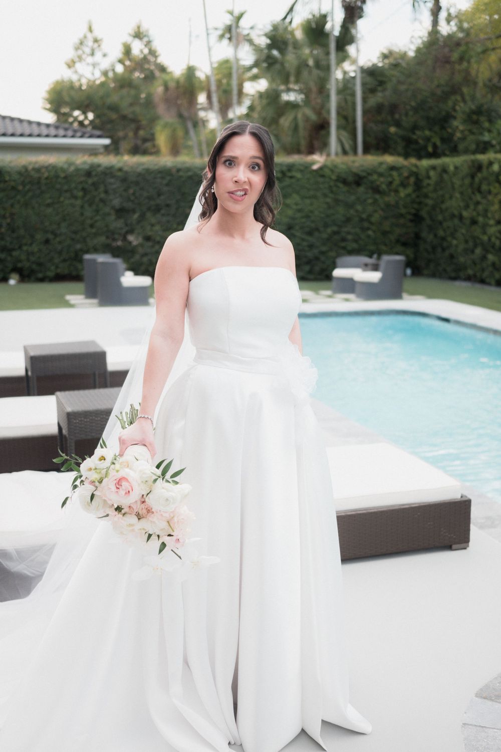 Bride in strapless white gown holding pink floral bouquet poses poolside at elegant outdoor wedding venue.