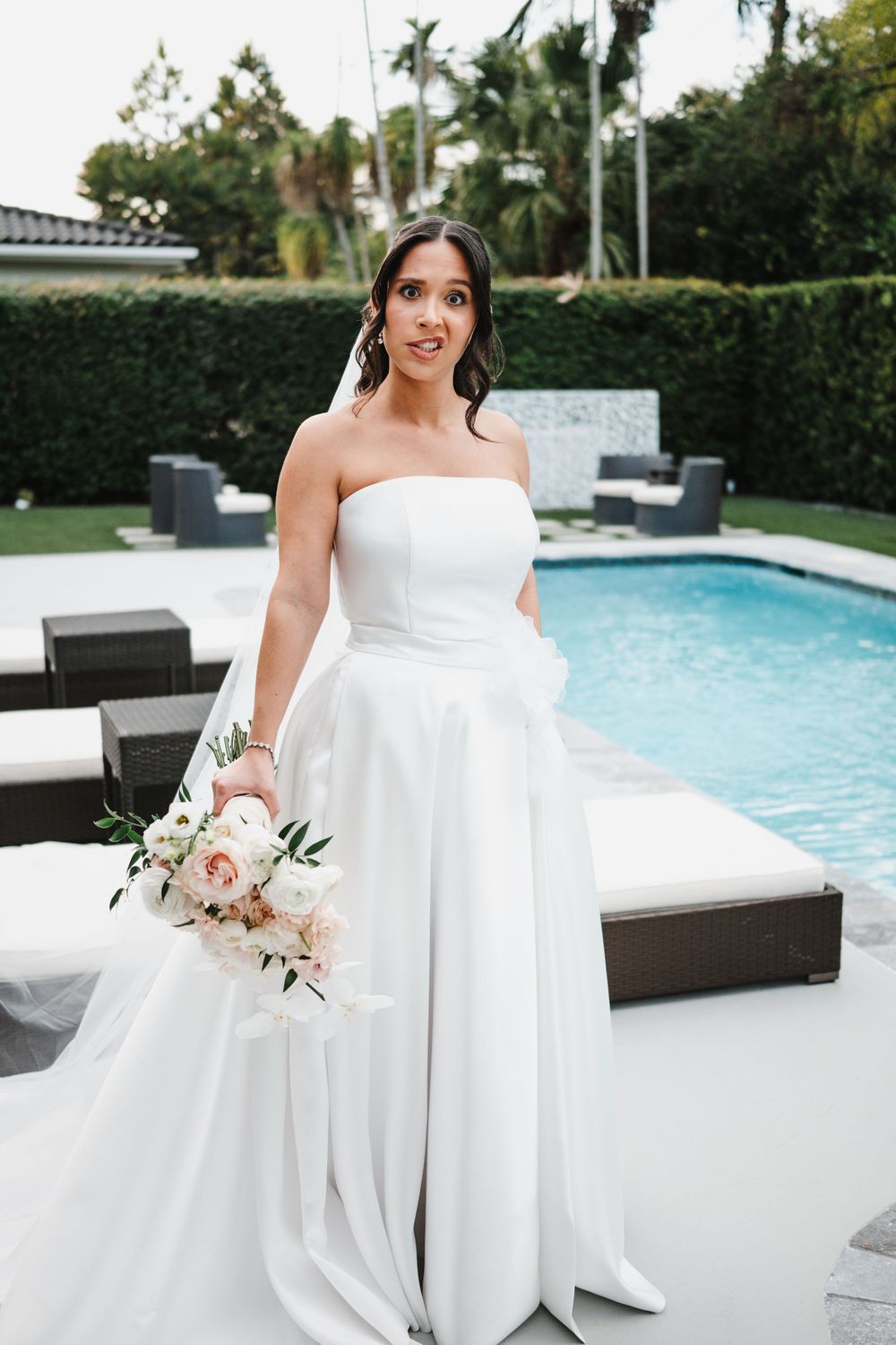 Elegant bride in a modern white wedding gown poses by a luxurious pool with pink and white floral bouquet.
