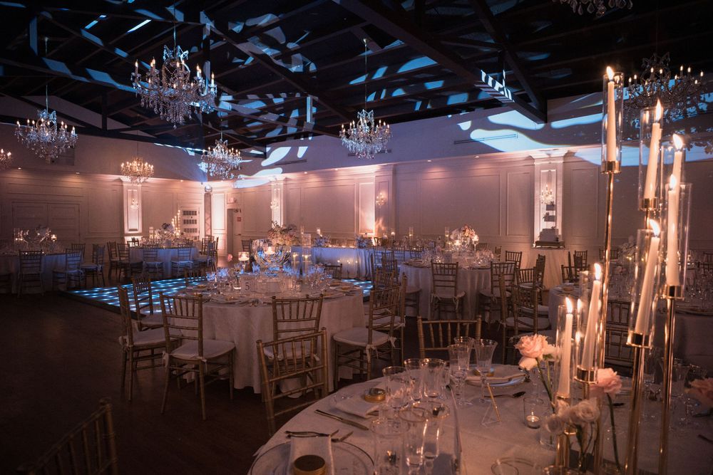 Elegant wedding reception hall with chandeliers, round tables, white linens, tall candle centerpieces, and soft blue ambient lighting.
