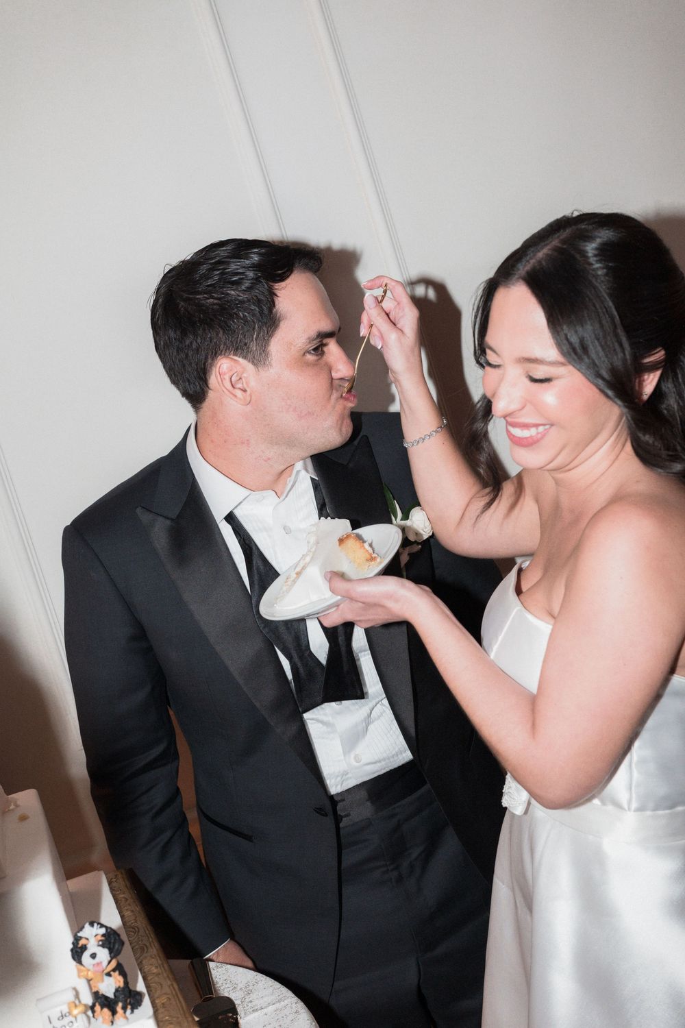 Bride feeding groom wedding cake at elegant reception, groom in black tuxedo, bride in white strapless gown, smiling.