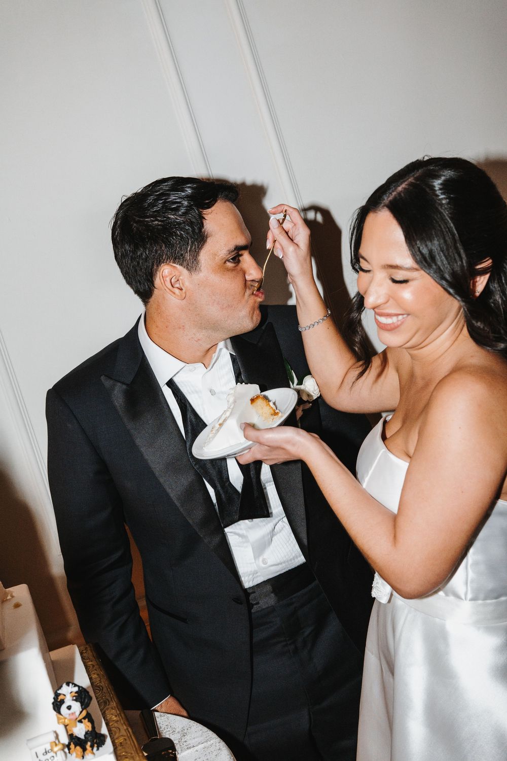 Playful couple in wedding attire share a sweet moment feeding each other cake during their reception celebration.