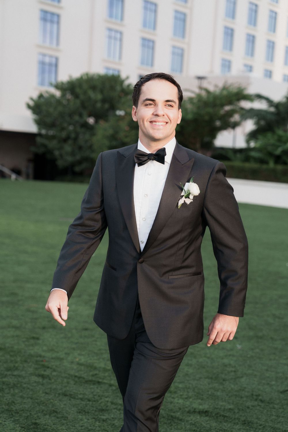 Smiling groom in black tuxedo with boutonniere walks on green lawn outside elegant venue.