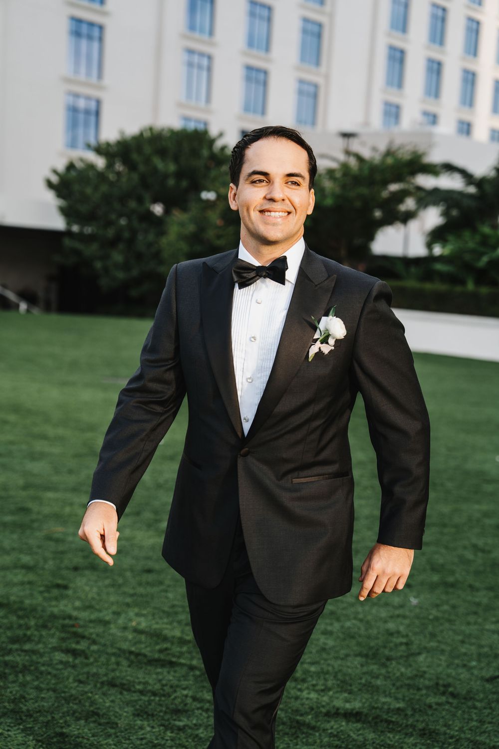A groom in a classic black tuxedo and bow tie poses on a green lawn outside an elegant building.