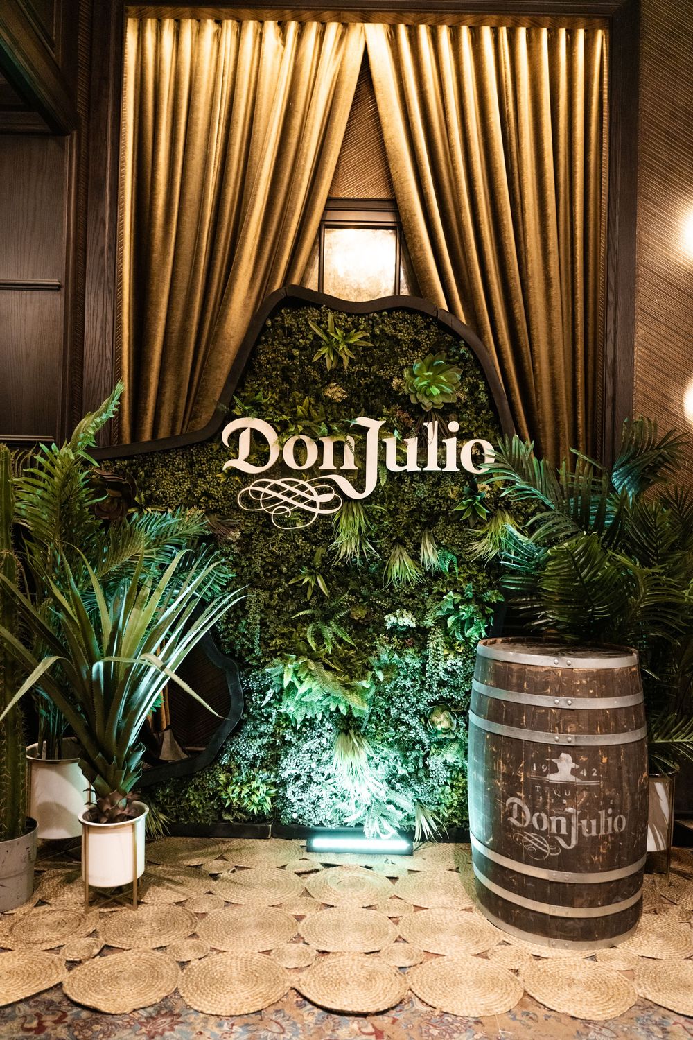 Decorative Don Julio tequila barrel display with dramatic lighting and tropical plants against gold curtains at an upscale venue.