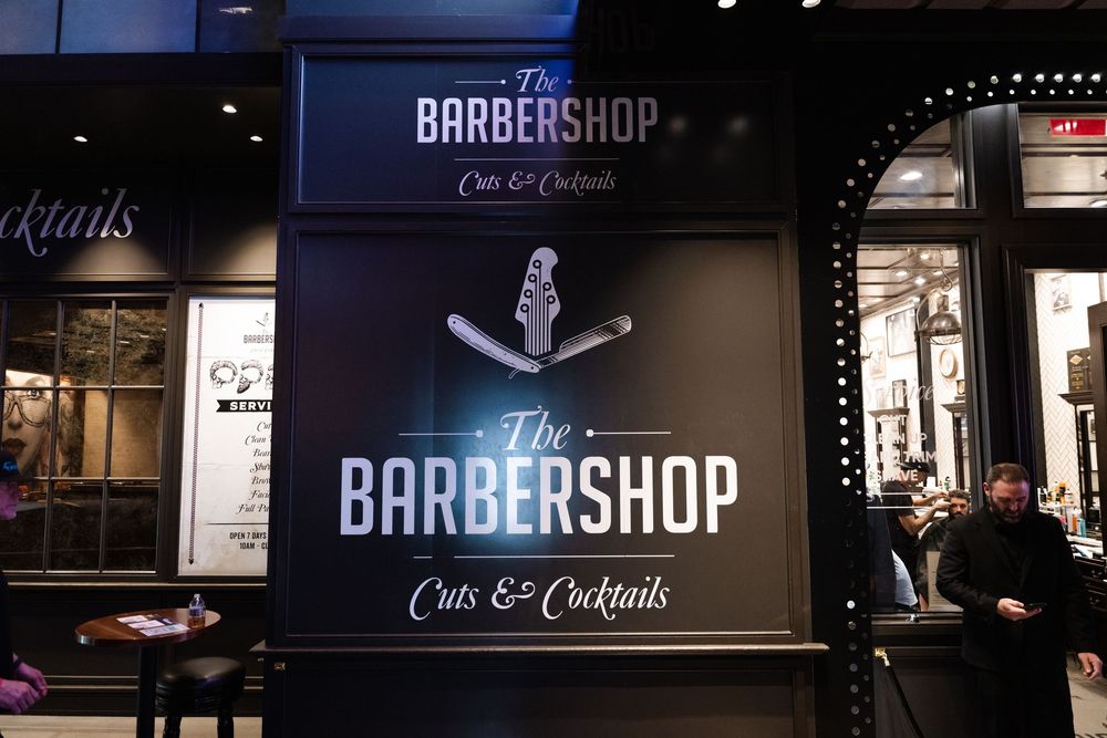 Illuminated storefront sign for 'The Barbershop Cuts & Cocktails' with black background and white lettering at night.